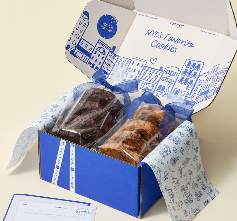An open blue and white gift box containing two individually wrapped cookies, with printed blue illustrations on the inside lid and tissue paper lining the box. A small card is visible in the lower left corner.